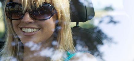A happy young woman wearing sunglasses looks out crystal-clear windows as she drives her car on a sunny day.
